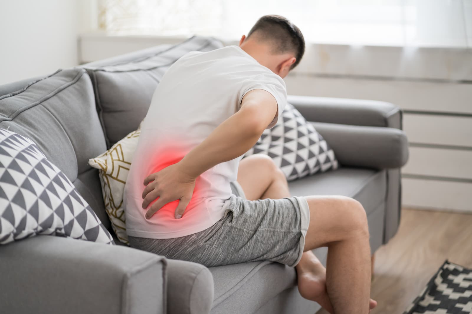 A man sitting on a grey sofa clutching his lower back in pain, with a glowing red highlight indicating the source of the inflammation.