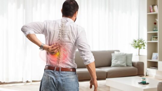 A man viewed from behind clutching his lower back, featuring a transparent overlay of a human spine with a glowing red and yellow area indicating lumbar pain.