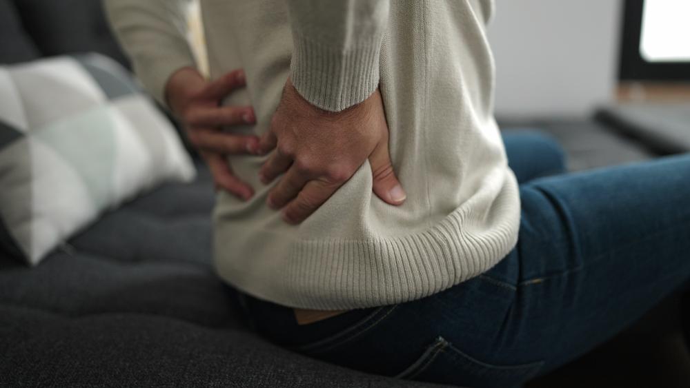Person sitting on a dark sofa, pressing both hands firmly against their lower back in pain.