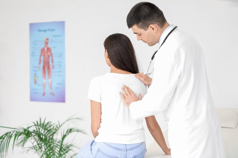 Doctor in a white coat examining the lower back of a seated female patient in a bright clinic room.
