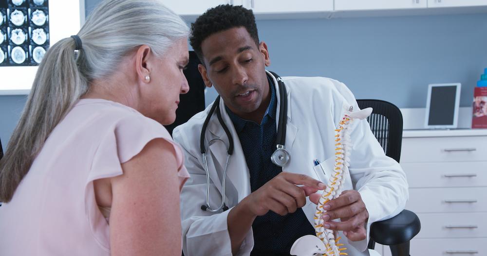 Doctor pointing at the neck area of a spine model while talking to an older female patient in a clinic.